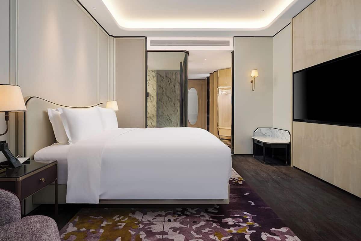 A modern hotel room with a large bed, bedside lamps, a TV, a small table, and a bathroom visible through an open door.