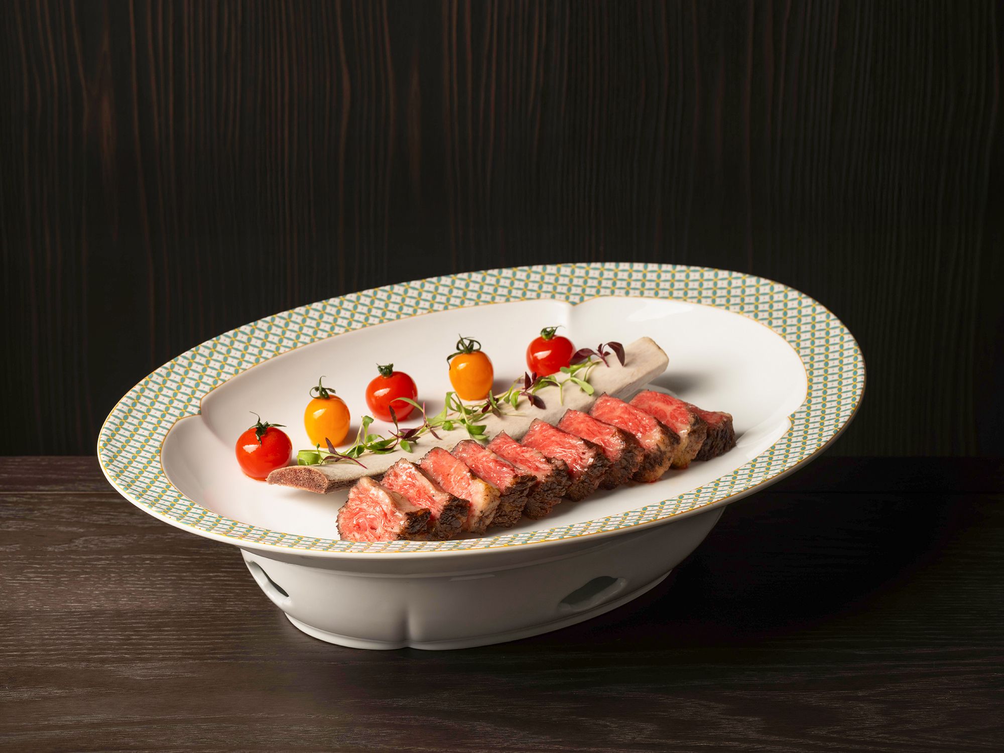 A white plate with a green and yellow pattern, containing sliced meat, cherry tomatoes, and greens.