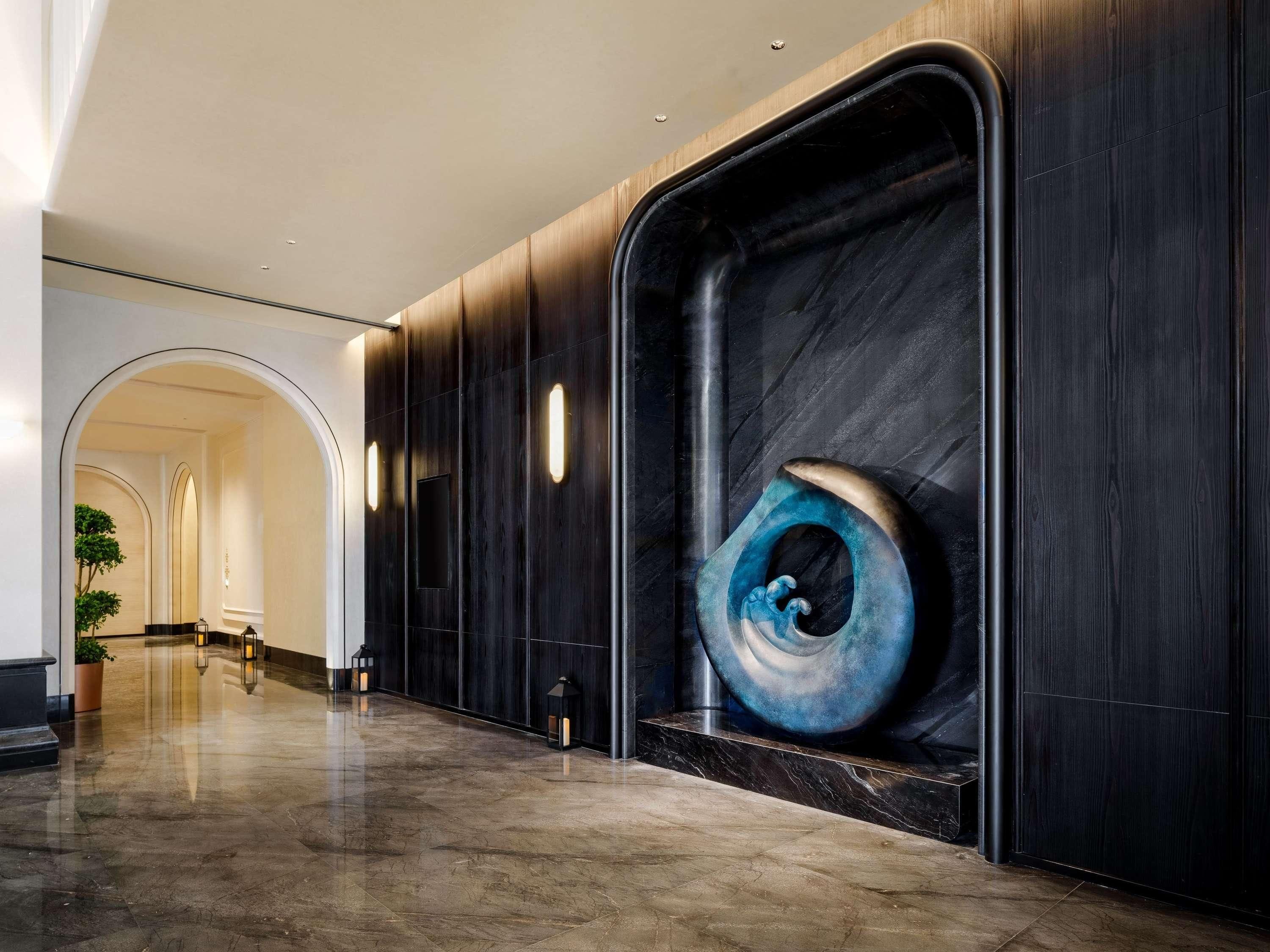 Un vestíbulo de hotel moderno con piso de mármol, paneles de madera oscura y una gran escultura abstracta.