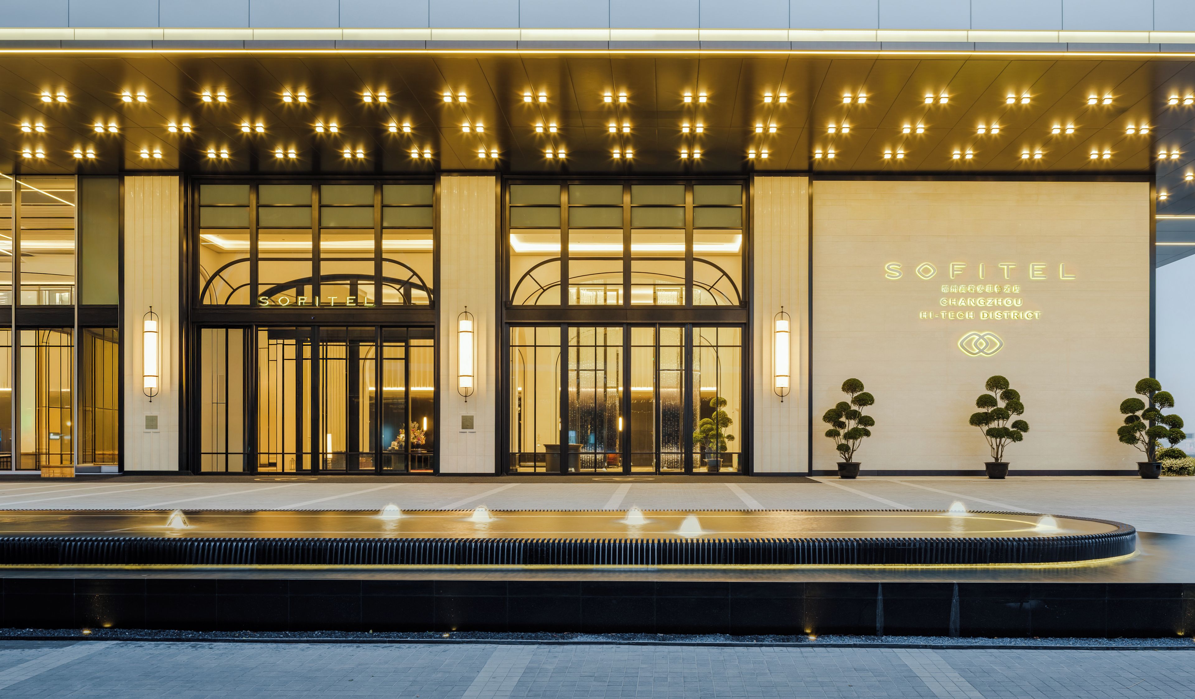 Entrada de hotel con diseño moderno, destacada por un amplio frente de vidrio, una fuente de agua y señalización iluminada.