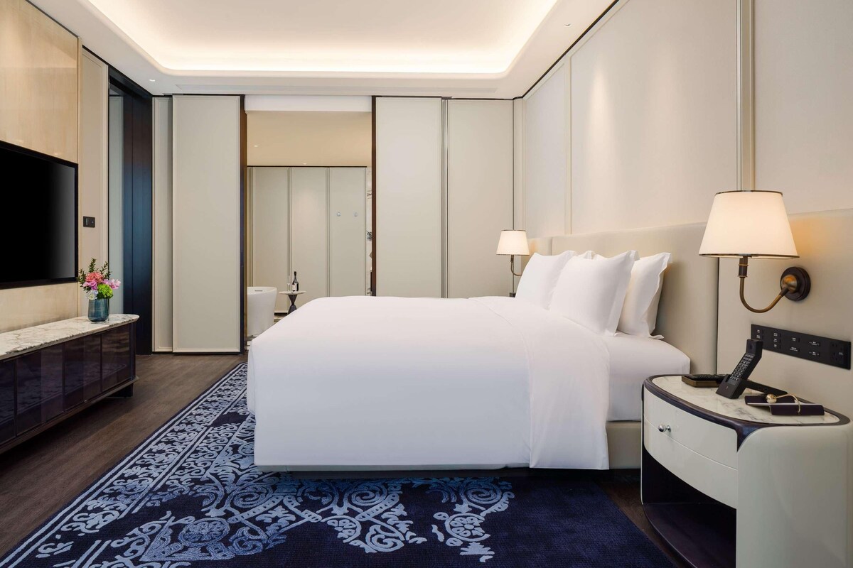 A modern hotel room with a large bed, a flat-screen TV, a bedside table with a lamp, and a desk with a telephone.