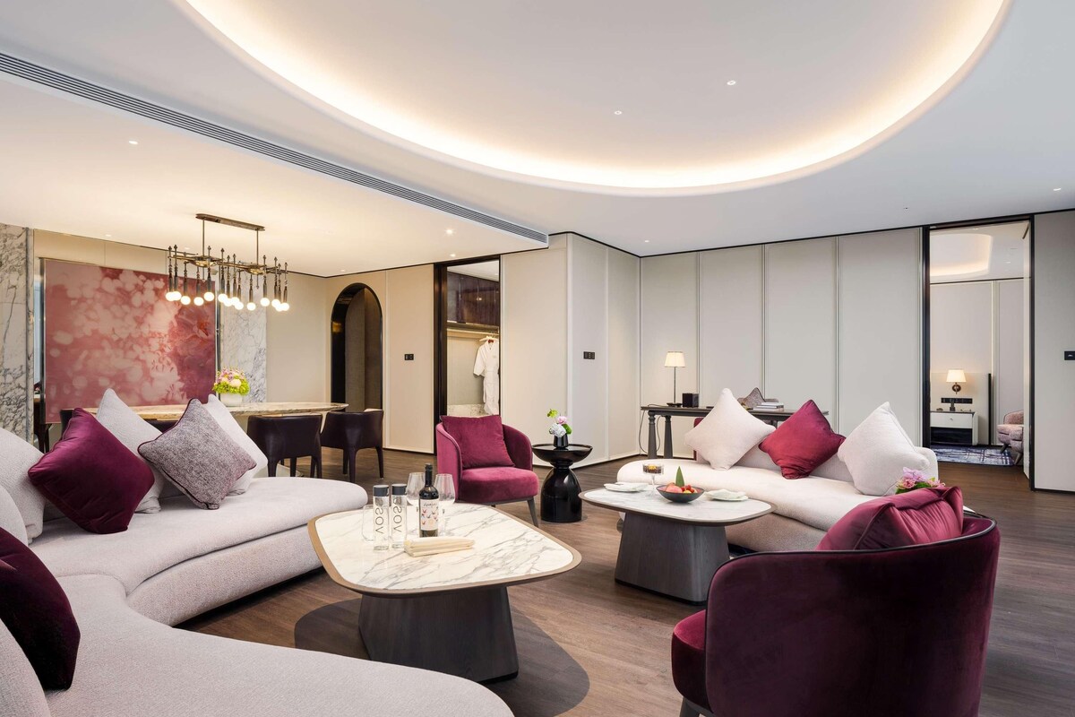 A modern hotel lobby with a curved ceiling, a seating area with sofas and armchairs, a dining table, and a large abstract painting.