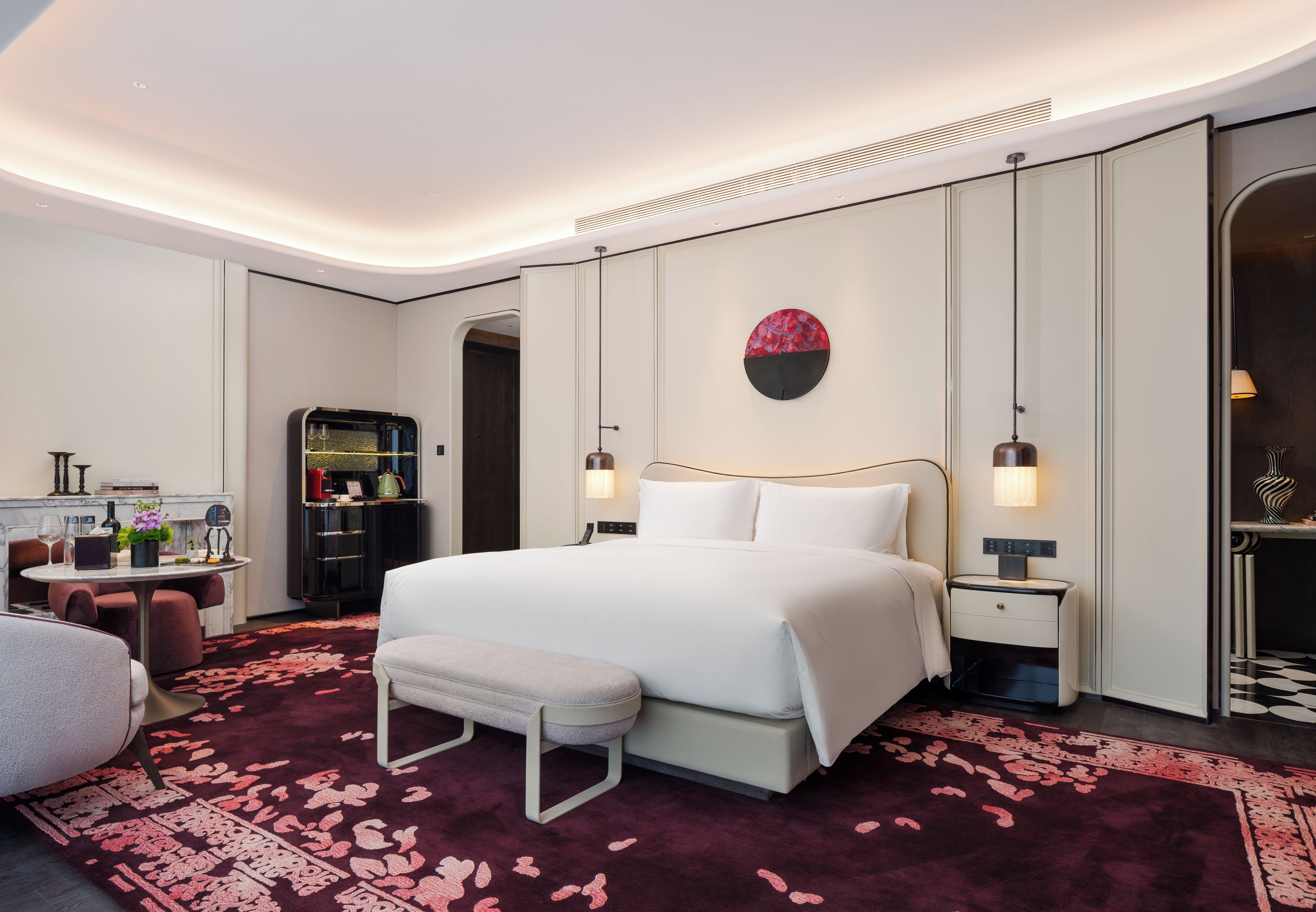 A modern hotel room with a large bed, bedside tables, a desk with a chair, a round mirror, and a decorative wall piece.