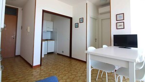 Ein Zimmer mit einem weißen Tisch, Stühlen, einem Fernseher, einer Tür und einer kleinen Küchenzeile.