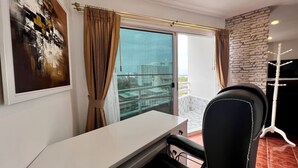 Una habitación moderna con un escritorio blanco, una silla negra, un ventanal con cortinas y vistas a la ciudad, además de una pared de piedra.