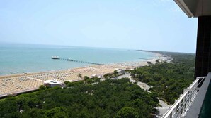 A beach with rows of sunbeds and umbrellas, a pier extending into the sea, and dense greenery in the foreground. - Holiday apartment mit Balkon und Meerblick (Lignano Sabbiadoro)