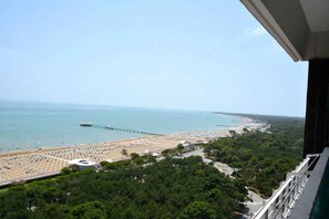 A beach with rows of sunbeds and umbrellas, a pier extending into the sea, and dense greenery in the foreground. - Holiday apartment mit Balkon und Meerblick (Lignano Sabbiadoro)