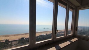 A room with large windows offering a view of a beach, a pier, and distant buildings. - Holiday apartment mit Balkon und Meerblick (Lignano Sabbiadoro)