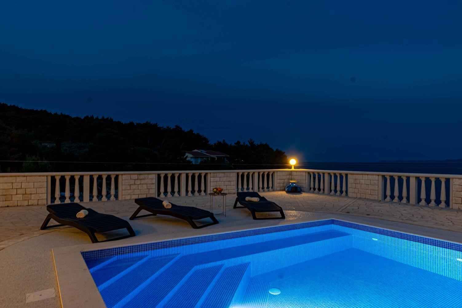 A swimming pool with lounge chairs and a lamp post, surrounded by a stone wall and overlooking a body of water.