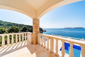 Ein Balkon mit Steinbalustrade, der Blick auf das klare, blaue Meer und eine hügelige Landschaft freigibt.