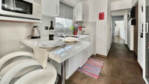 Eine moderne Küche mit weißen Schränken, einer Mikrowelle, einem Wasserkocher und einer Weinflasche. Es gibt einen Essbereich mit einem Tisch für zwei Personen, einem Teppich und einem Fenster mit Blick.