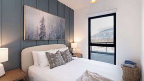 Ein Schlafzimmer mit Bett, Nachttisch, Lampe, einem Gemälde und einem Fenster mit Blick auf eine verschneite Landschaft.