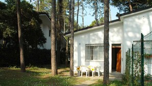 A white building with a door and window, surrounded by trees and a small garden with a table and chairs. - Holiday apartment mit Terrasse (Lignano Sabbiadoro)