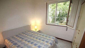 A bedroom with a bed, a nightstand with a lamp, a window with a view of greenery, and a door with a curtain. - Holiday apartment mit Terrasse (Lignano Sabbiadoro)