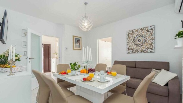 Ein moderner Essbereich mit einem weißen Tisch, gedeckt für vier Personen, umgeben von beigefarbenen Stühlen. Im Hintergrund steht ein braunes Sofa und über dem Tisch befindet sich ein dekoratives Wandbild.