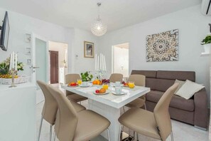 Un espace repas moderne avec une table blanche dressée pour quatre personnes, entourée de chaises de couleur beige. On aperçoit un canapé marron à l’arrière-plan et un élément décoratif mural au-dessus de la table.