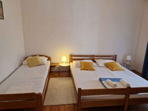 Ein Zimmer mit zwei Holzbetten, jeweils mit weißer Bettwäsche und gelben Kissen. Dazwischen steht ein kleiner runder Tisch mit einer Lampe.