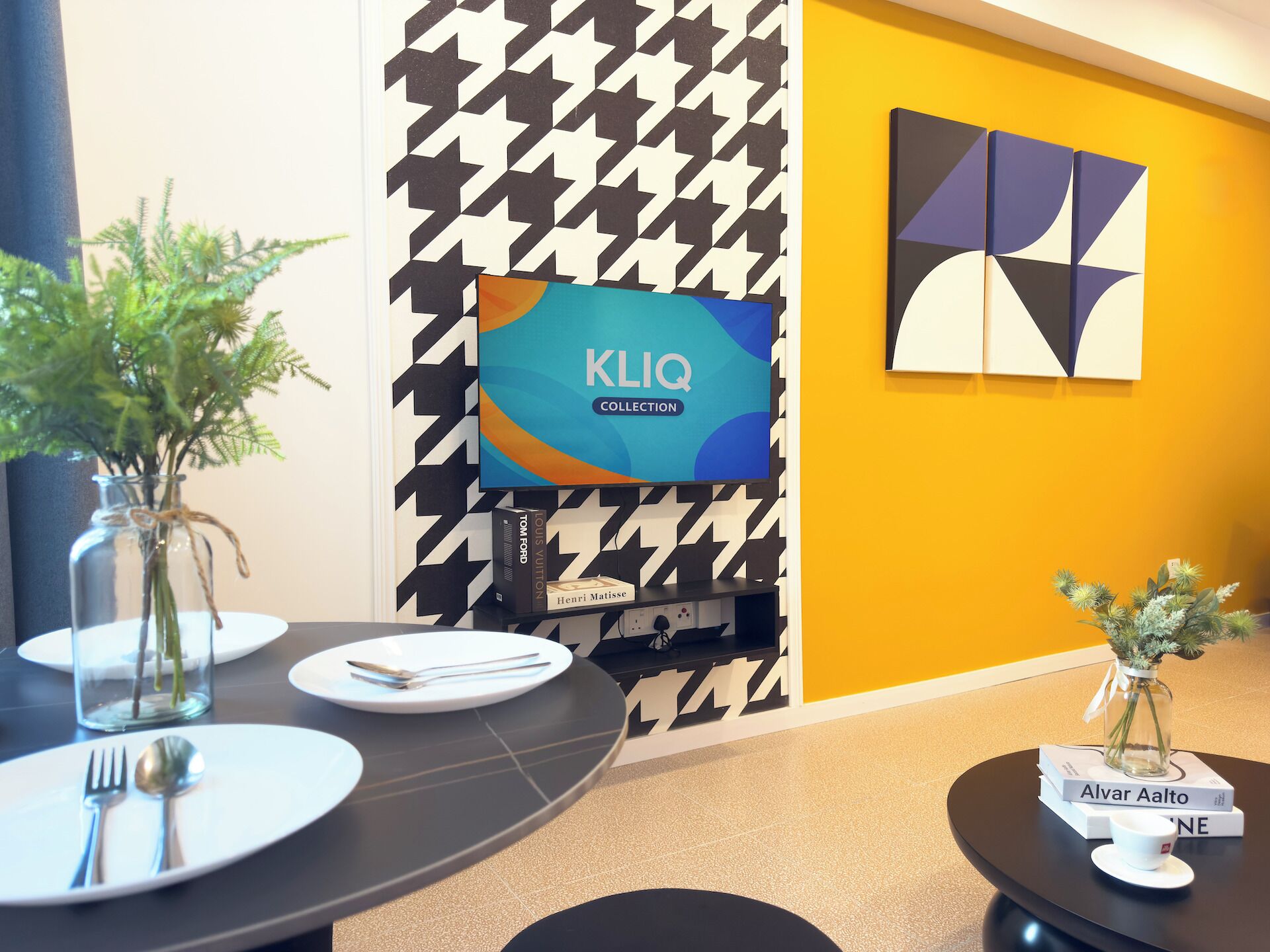 Une salle de conférence moderne avec un mur orné d’un motif à carreaux, un écran de télévision de la collection KLIQ et une table agrémentée d’un vase de plantes et d’un livre intitulé « Alvar Aalto ».