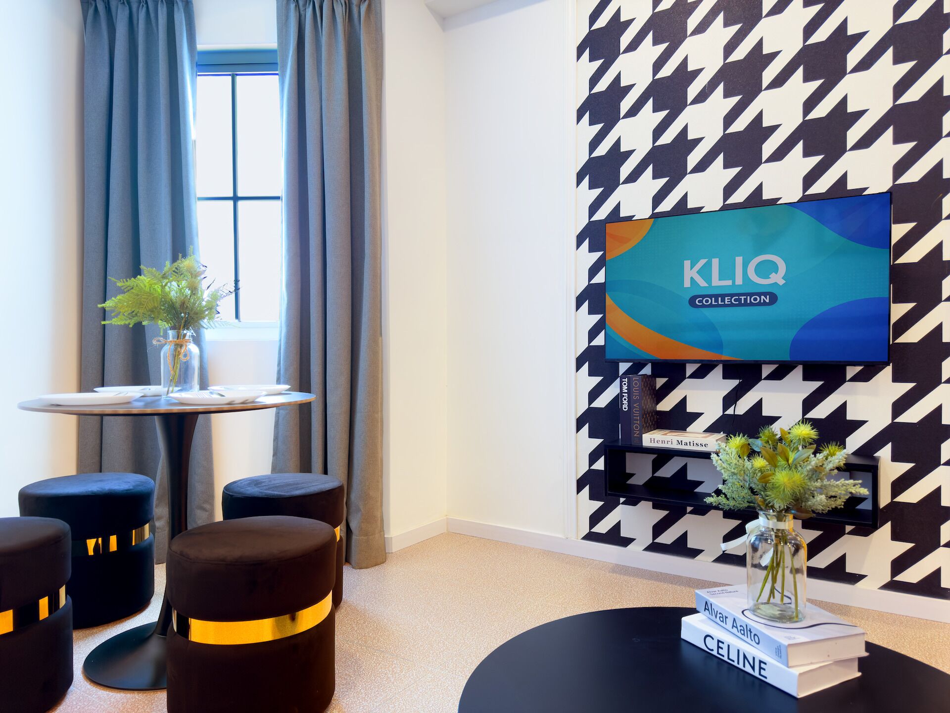Una habitación de hotel moderna con un estampado a cuadros en blanco y negro tipo pata de perro, una mesa redonda con un jarrón de plantas verdes y otomanas negras con detalles dorados.
