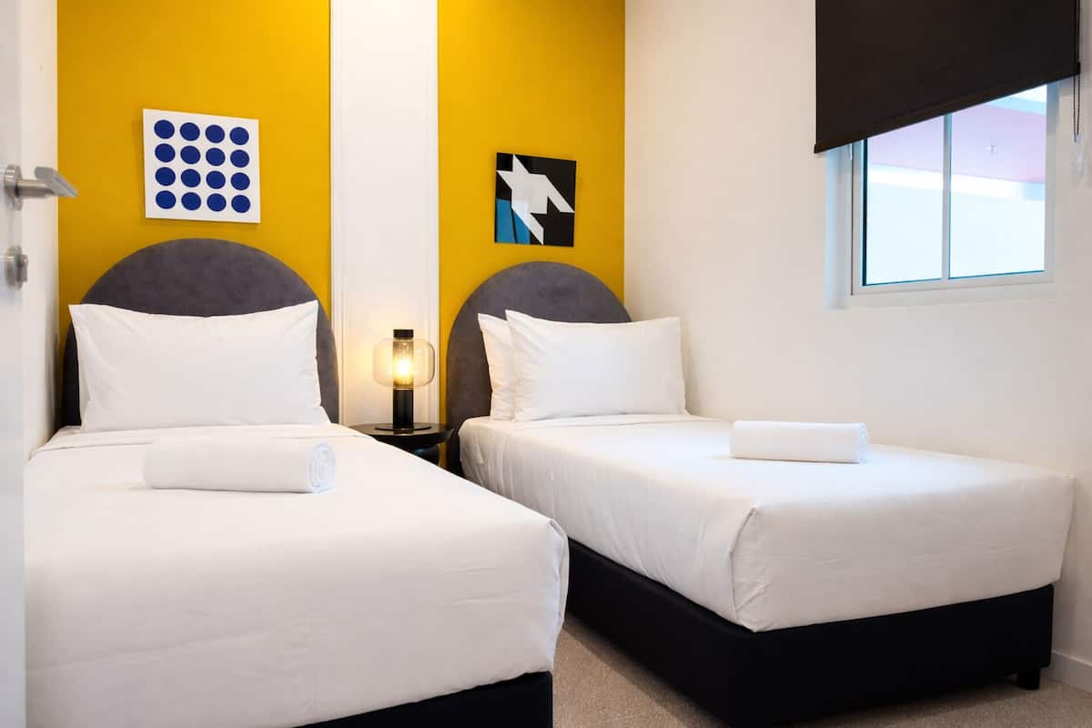 A hotel room with two beds, a nightstand with a lamp, a wall with a geometric art piece, and a window with a view.