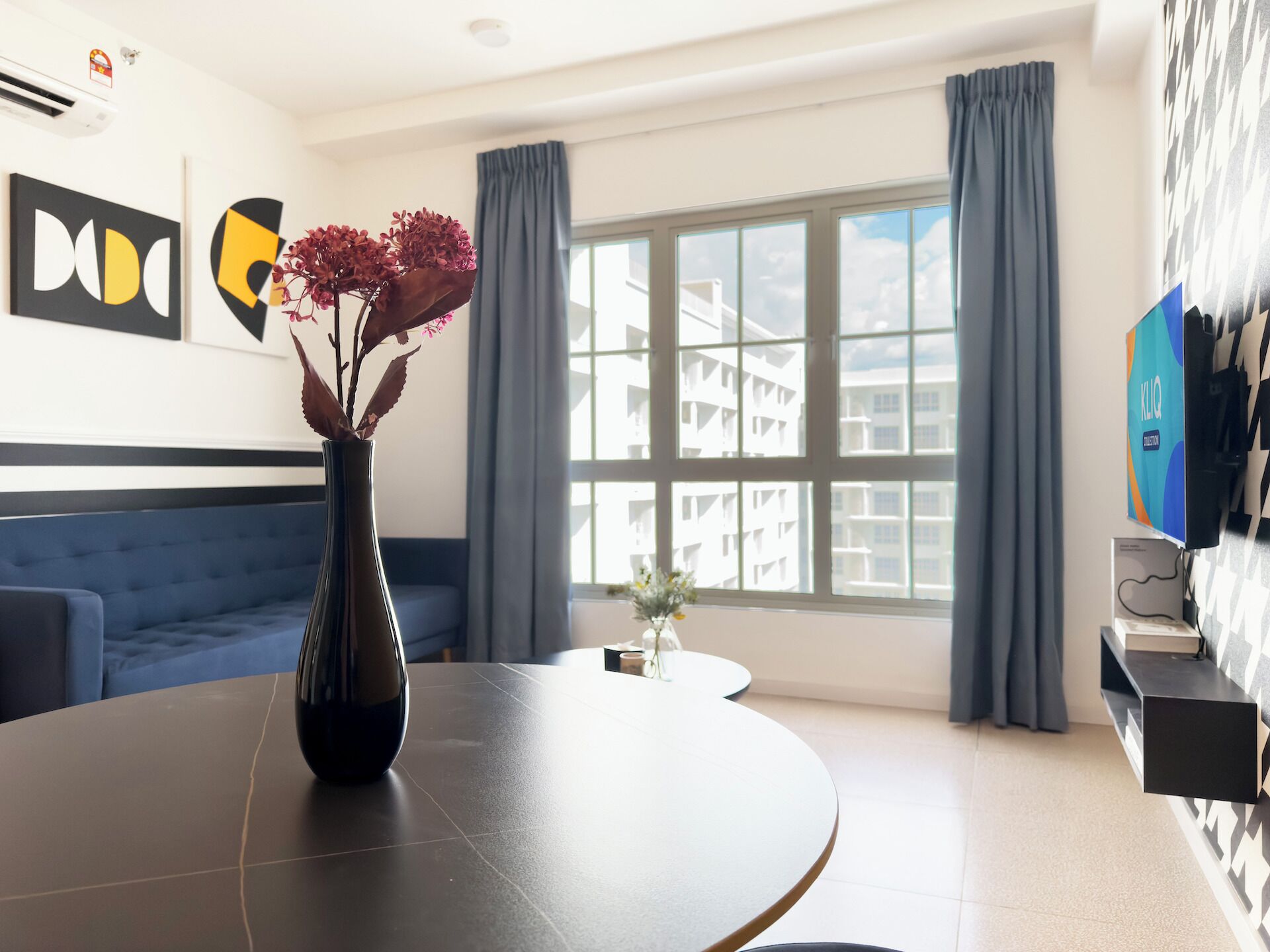 A modern hotel room with a round dining table, a blue sofa, a large window with curtains, and a flat-screen TV mounted on the wall.