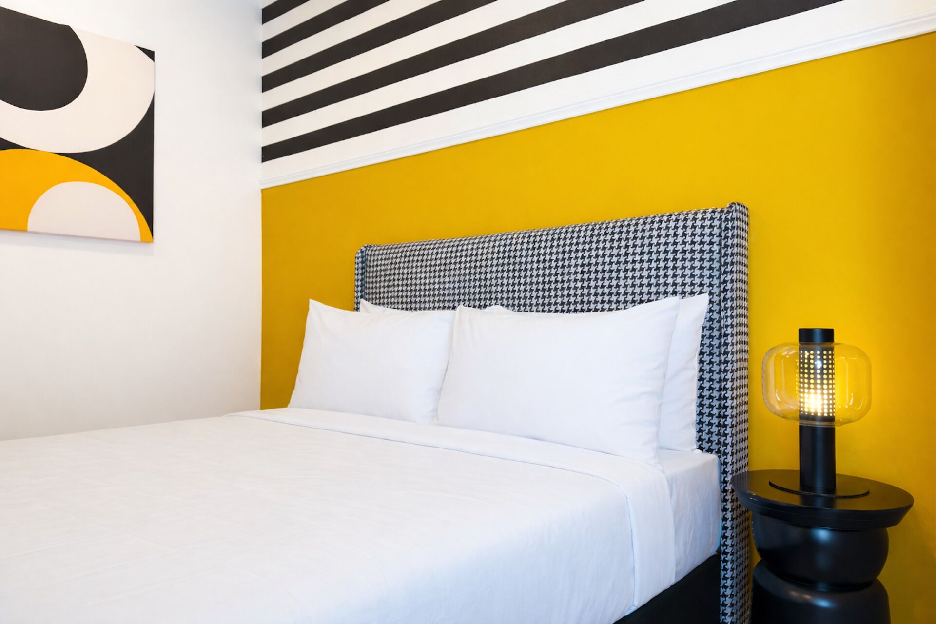 Ein Bett mit weißer Bettwäsche, ein Kopfseitenverkleidung mit Hunde-Zahn-Muster und eine schwarze Lampe auf einem schwarzen Nachttisch daneben.