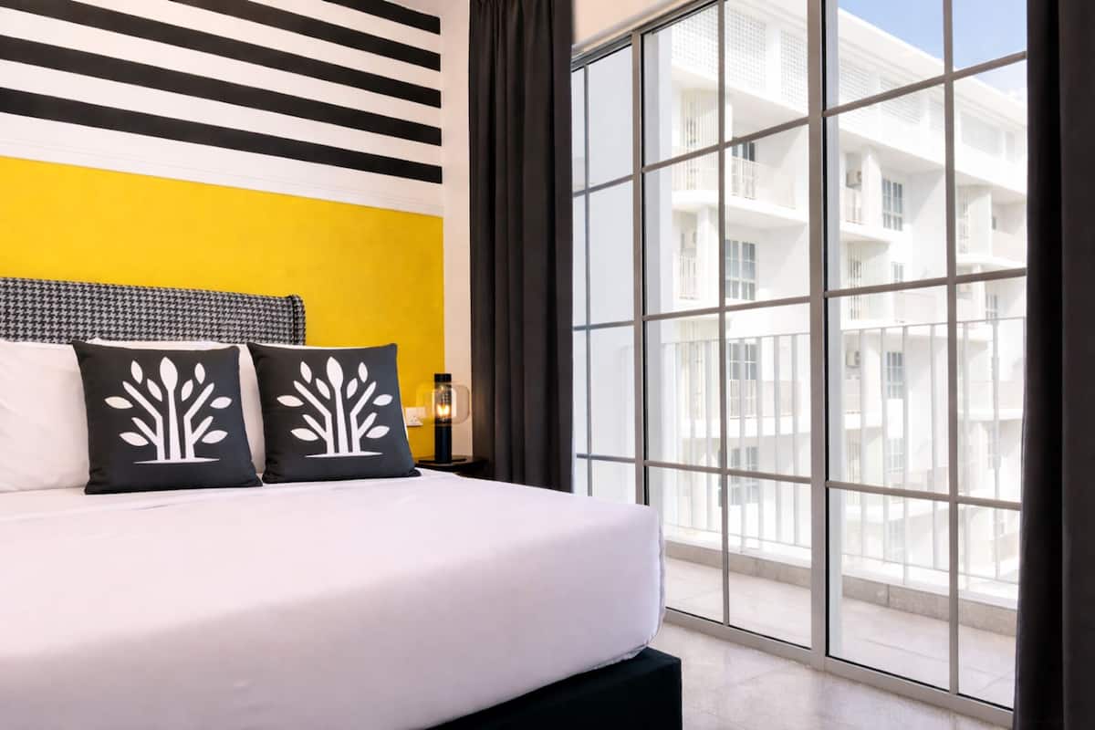 A hotel room with a large bed, two black and white tree-patterned pillows, a houndstooth headboard, and a view of a white building through floor-to-ceiling windows.