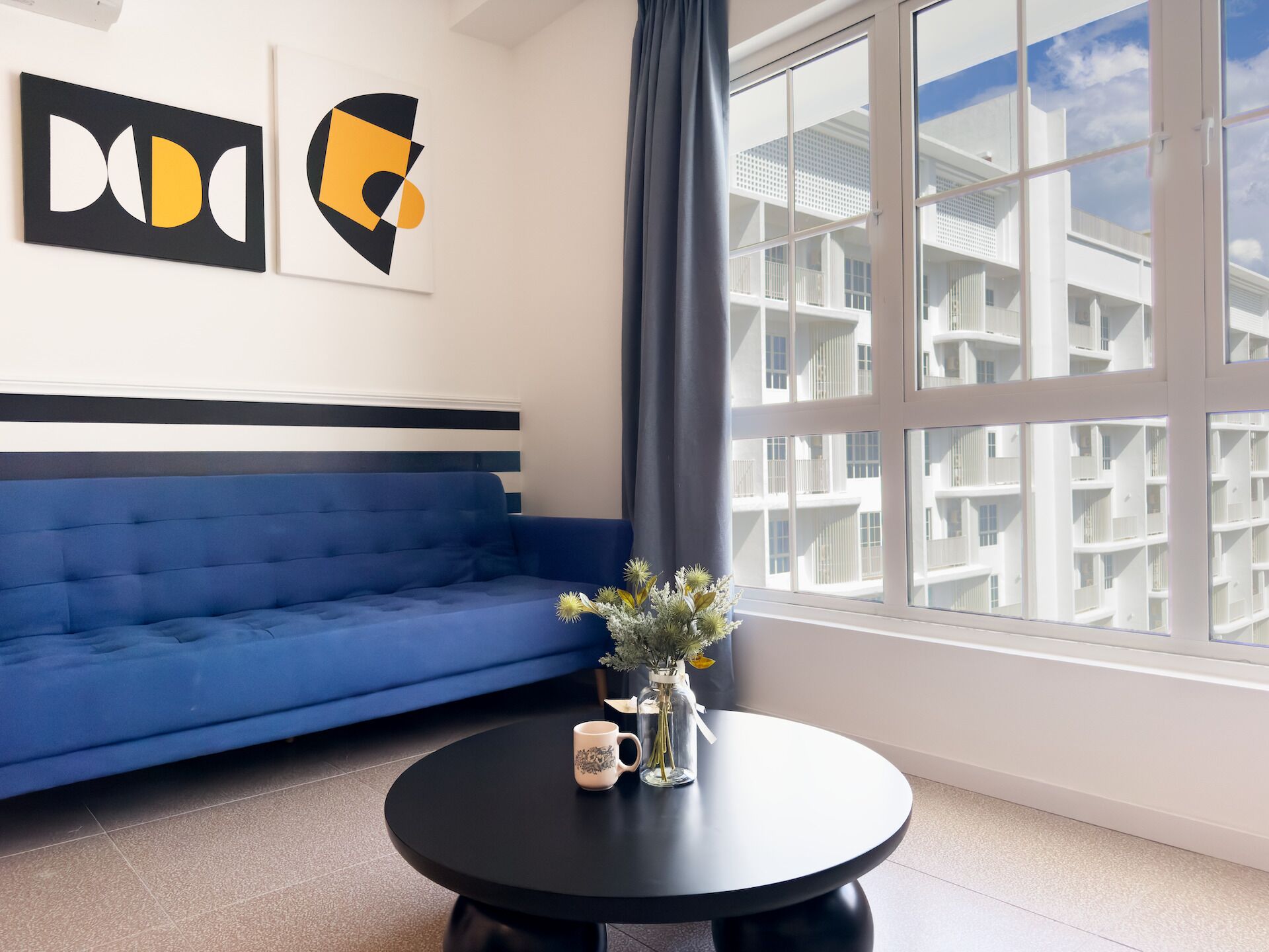 Ett modernt vardagsrum med en blå soffa, ett runt svart bord med en vas och två muggar, och ett stort fönster med utsikt över en vit byggnad.