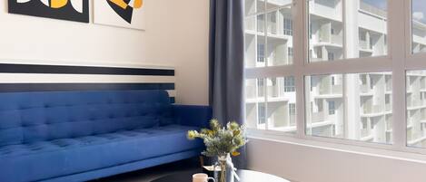 A modern living room with a blue sofa, a round black table with a vase and two mugs, and a large window with a view of a white building.