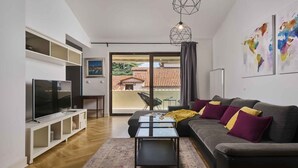 Uma sala de estar moderna com uma televisão de tela plana, um sofá com almofadas, uma mesa de centro e vista para um edifício com telhado de cerâmica.