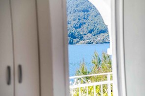 Blick durch ein Fenster mit weißer Brüstung auf ein Gewässer und einen bewaldeten Hügel.