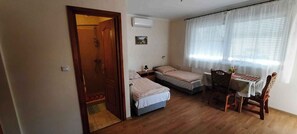 A small, well-lit room with two single beds, a wooden dining table with chairs, a small table with a vase, and a shower area visible through an open door. - Holiday apartment with air conditioning (Keszthely)