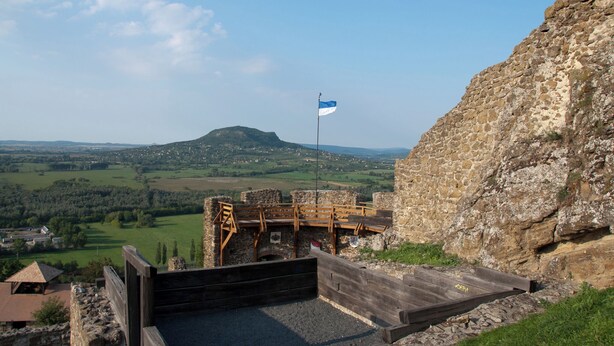 Ein Steingebäude mit einer Flagge, einer Holzplattform und Blick auf eine grüne Landschaft.