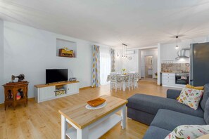 Ein modernes Wohnzimmer mit einem Flachbildfernseher, einem Holztisch und einem grauen Sofa. Es gibt einen Essbereich mit Tisch und Stühlen sowie eine Küchenzeile mit Kühlschrank und Schränken.