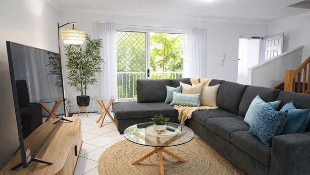 Un salon moderne avec un canapé gris, une table basse en verre, un téléviseur posé sur un meuble en bois, une plante en pot et un balcon donnant sur un paysage verdoyant.
