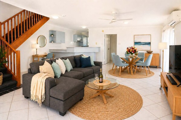 A modern living room with a grey sectional sofa, a coffee table with a bottle and glasses, a dining area with a round table and chairs, and a kitchen in the background.