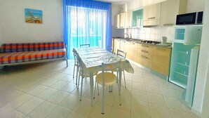 Un espacio habitable compacto con una mesa de comedor, sillas, un sofá, una cocina con armarios y electrodomésticos, y un gran ventanal con cortinas.