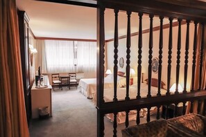 Een hotelkamer met een bed, een bureau en een stoel. Er is een raam met gordijnen, een lamp op het bureau en een fotolijst aan de muur.