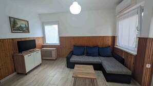 A living room with a wooden panel wall, a grey sectional sofa with blue cushions, a wooden coffee table, a TV on a wooden cabinet, and a window with a white blind. - Holiday apartment with WLAN and air conditioning (Zamárdi)