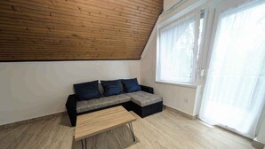 Ein modernes Schlafzimmer im Loft-Stil mit schrägverdeckter Decke, einem grauen Ecksofa, einem Holztisch und einem großen Fenster mit durchscheinenden Vorhängen.