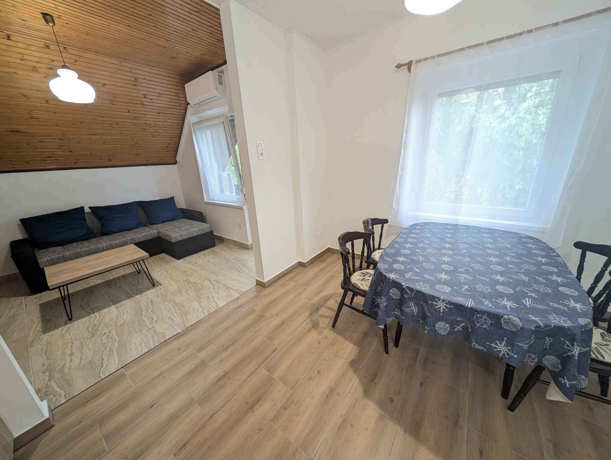 A room with a wooden ceiling, a sofa with cushions, a dining table with chairs, and a window with curtains.