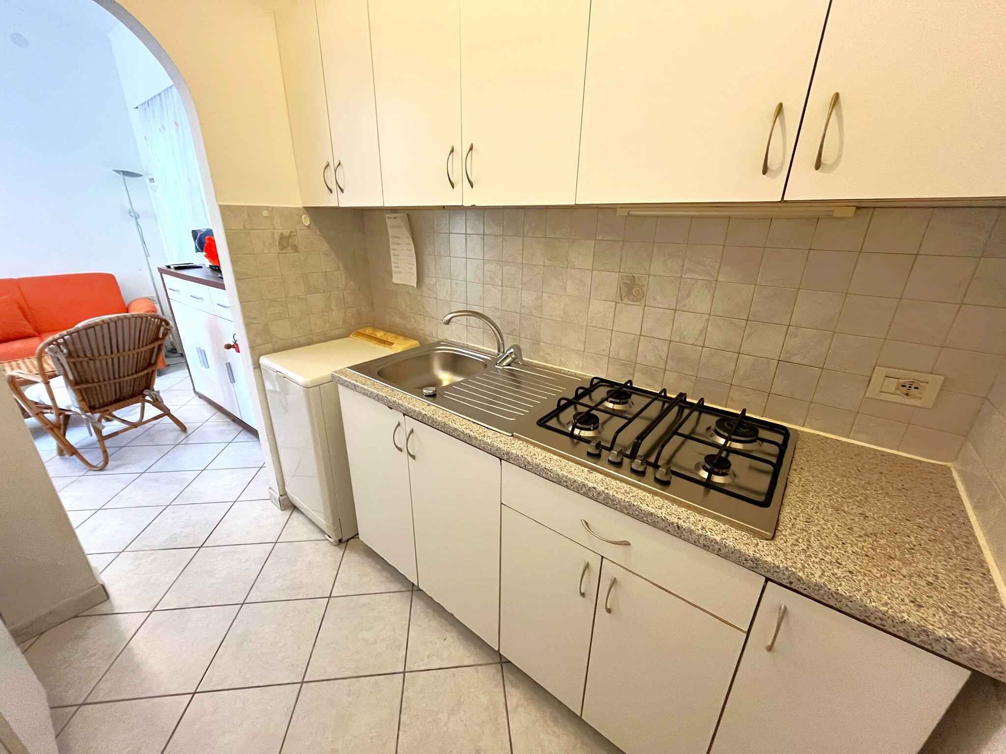 Una cocina compacta con gabinetes blancos, estufa a gas y un lavabo. Hay una pequeña encimera y una silla en la zona de estar contigua.
