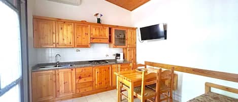 A kitchen with wooden cabinets, a dining table with chairs, a TV mounted on the wall, and a window.