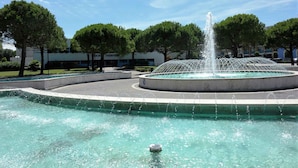 Una fontana con getti d'acqua in un laghetto circolare, circondata da alberi ed edifici.
