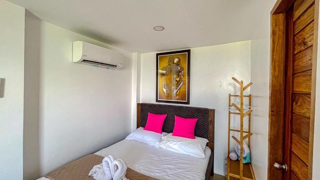 A hotel room with a bed, white bedding, two pink pillows, a wooden headboard, a wall-mounted air conditioner, a framed artwork, and a wooden ladder leaning against the wall.