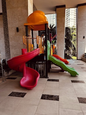 A children's play area with slides and a climbing structure, set against a backdrop of tall buildings and a tiled floor. - Cozy 1-bedroom apartment in awesome Pattaya with AC and gym access (pattaya)