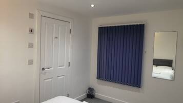 A hotel room with a large bed, a door with a handle, a window with blinds, a small trash bin, and a wall-mounted mirror.