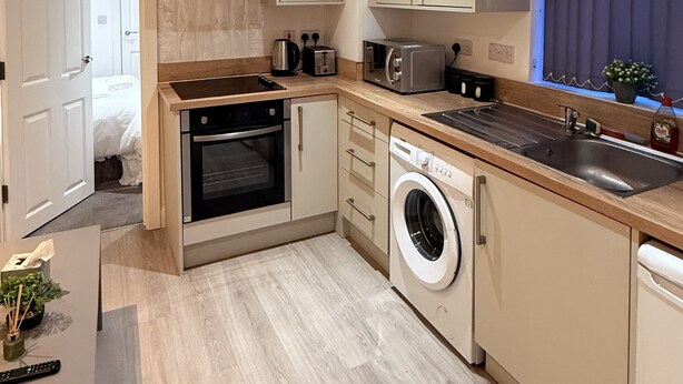 Cocina compacta con horno integrado, microondas y lavadora, con suelo de madera clara y muebles de tonos neutros.