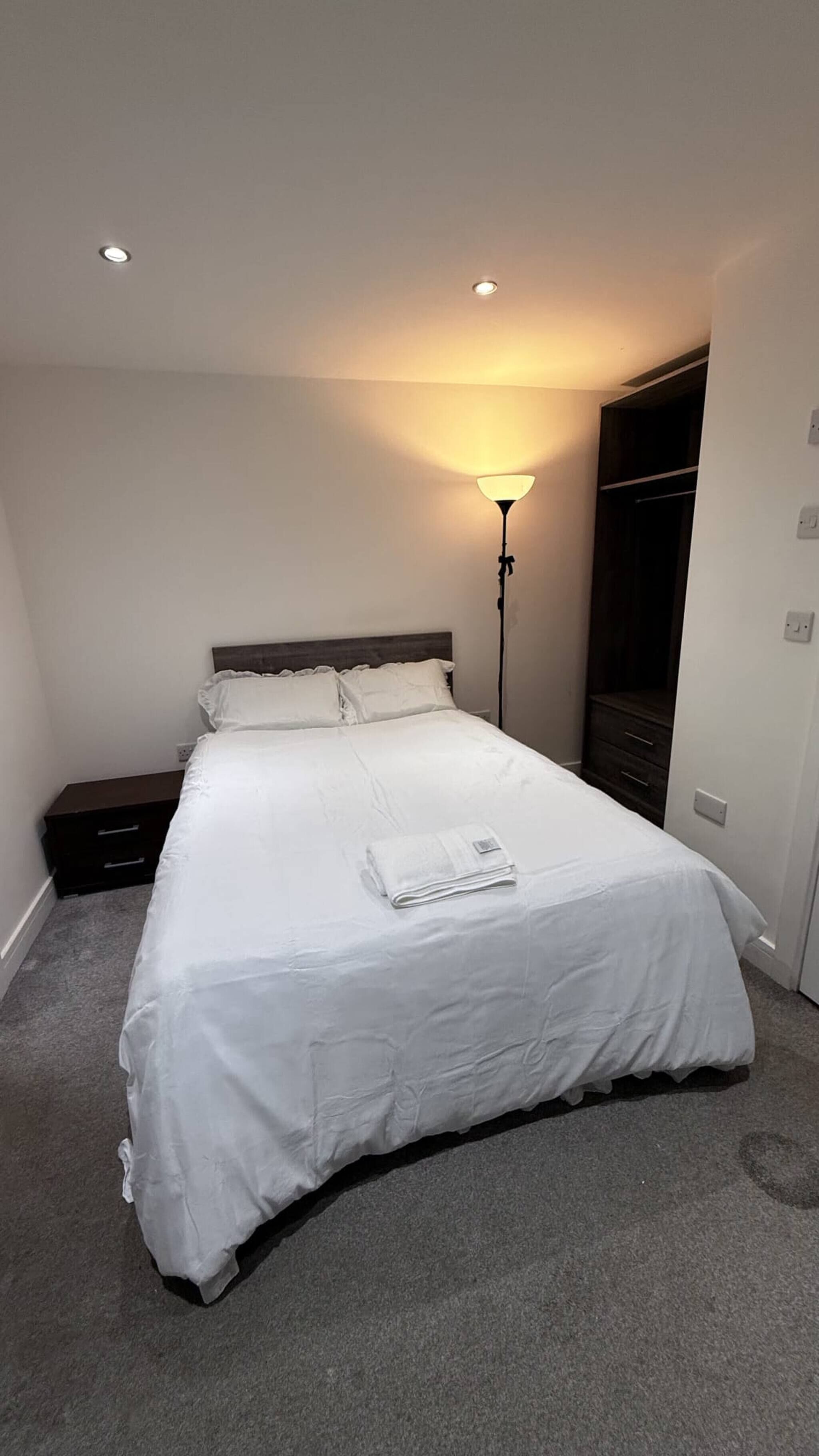 Ein ordentlich bezogenes Bett mit weißer Decke und Kissen, ein Nachttisch, eine Stehlampe und ein dunkler Holzschrank.