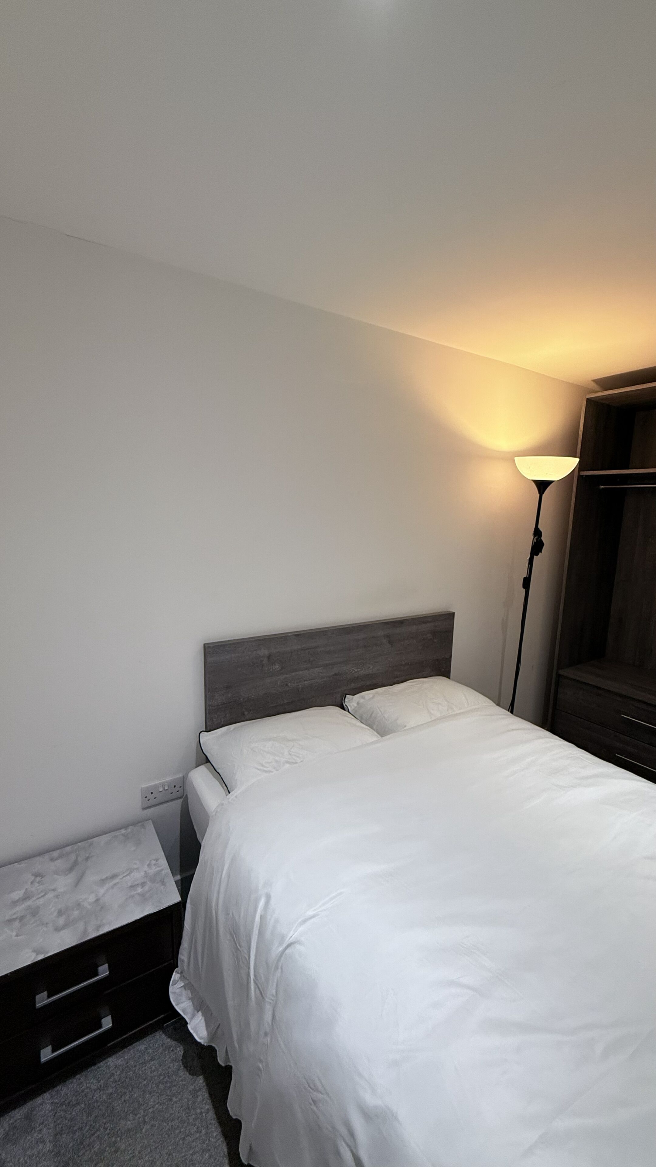 A neatly made bed with white linens, a bedside table with drawers, a floor lamp, and a wooden headboard.
